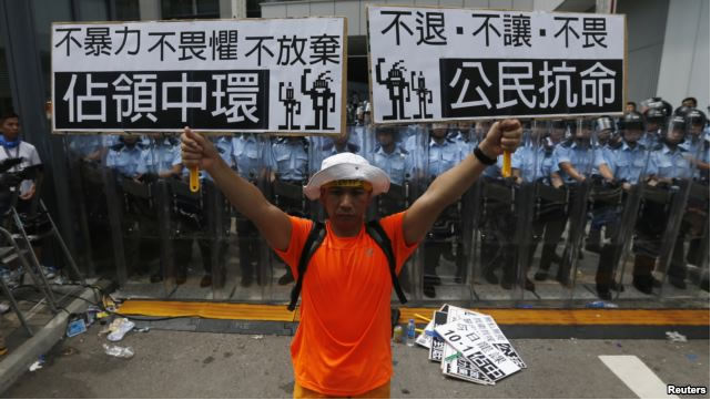 Một người biểu tình cầm áp phích 'Chiếm lĩnh Trung tâm' (trái) và 'Bất tuân Dân sự' đứng trước lực lượng cảnh sát chống bạo động bên ngoài trụ sở chính phủ ở Hồng Kông, 27/9/2014
